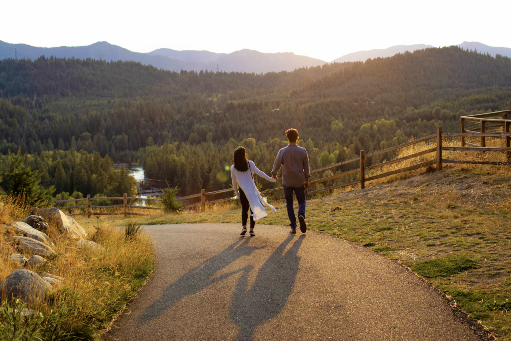 A couple walks hand in hand down a path with a scenic view of mountains, trees, and a setting sun.